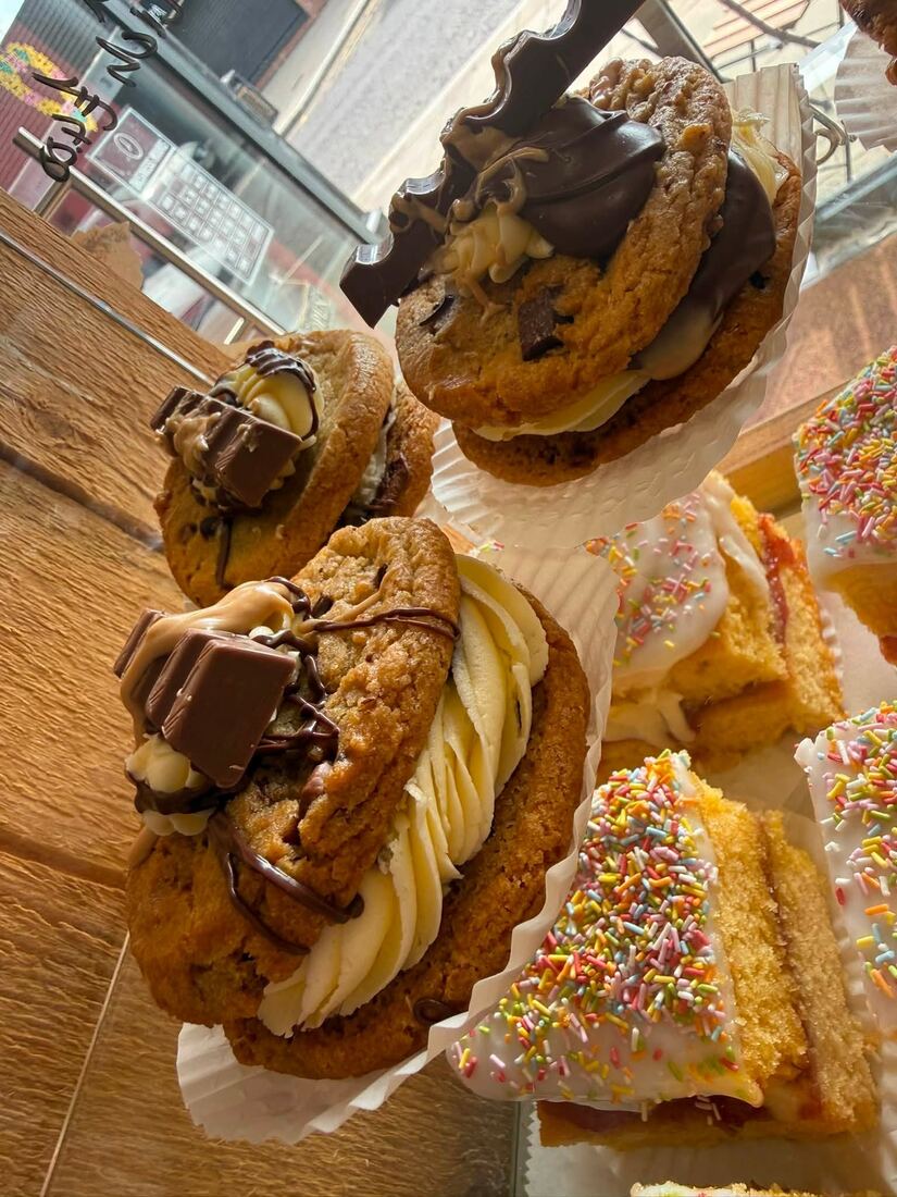 Loaded cookie sandwiches piped with buttercream and topped with chocolate, beside iced sprinkle traybakes